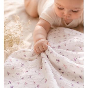 Könnte beinhalten: Ein Baby greift nach einer weißen Decke mit einem zarten Blumenmuster in Lila- und Rosatönen. Die Decke ist weich und strukturiert. Der Text oben lautet "weich für empfindliche Babyhaut."