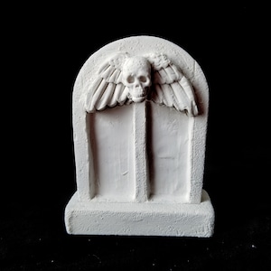 以下が含まれることがあります： 上部に頭蓋骨と翼が彫られた白い装飾墓石。墓石は長方形で、上部が丸く、表面はざらざらしています。背景は黒です。