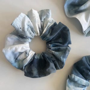 Puede incluir: Un scrunchie de tejido azul y blanco tie-dye. El scrunchie está hecho de dos tejidos diferentes, uno con fondo blanco y el otro con fondo azul. El scrunchie está anudado y listo para ser usado.