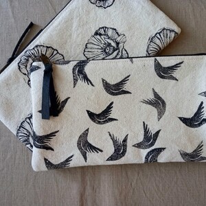May include: Two canvas zipper pouches. The larger pouch features a pattern of black flying birds, while the smaller pouch has a floral design. Both pouches have a natural, off-white background and black zippers.