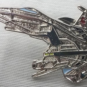 May include: A silver-colored metal pin of a futuristic spaceship with green, red, and blue accents.
