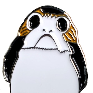 May include: A white, black, and orange enamel pin of a character from Star Wars. The character has large eyes, a small mouth, and two small legs.