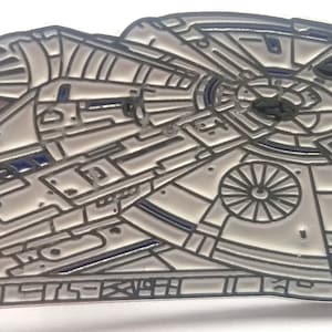 May include: A silver metal pin of the Millennium Falcon spaceship from Star Wars. The pin is detailed with black lines and blue accents.