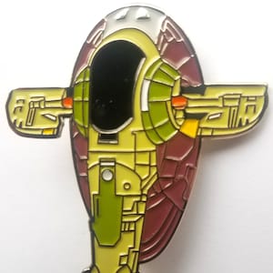 May include: A yellow, green, and brown enamel pin of a Star Wars spaceship, the Slave I.