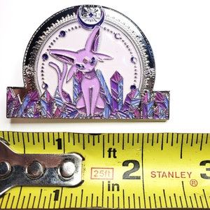May include: A purple and silver enamel pin featuring a pink Eeveelution Pokemon with a crescent moon and stars above it. The Pokemon is standing on a bed of purple and blue crystals.