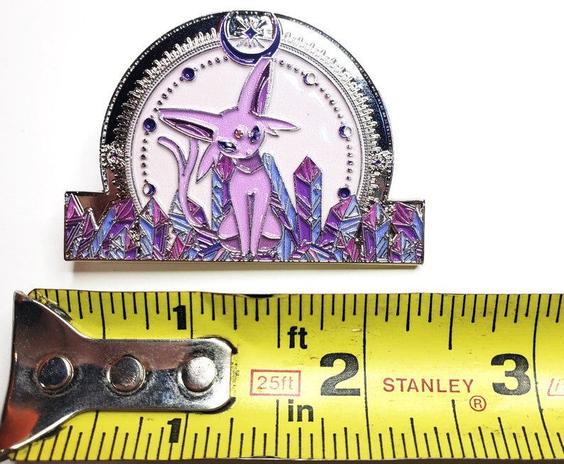 May include: A purple and silver enamel pin featuring a pink Eeveelution Pokemon with a crescent moon and stars above it. The Pokemon is standing on a bed of purple and blue crystals.