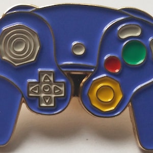 May include: A purple and gold GameCube controller pin. The controller has a circular button in the center, a directional pad, and four face buttons. The face buttons are red, green, yellow, and white.