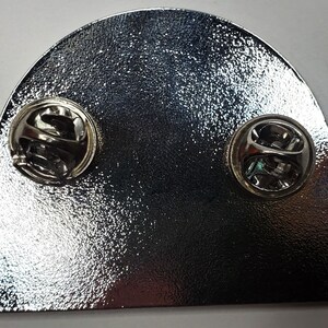 May include: The back of a silver metal pin with two pin backs. The pin is shaped like a half circle with a flat base.