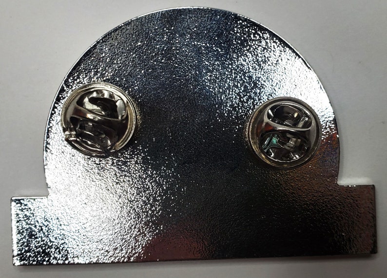 May include: The back of a silver metal pin with two pin backs. The pin is shaped like a half circle with a flat base.