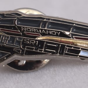 May include: A silver metal pin shaped like a spaceship with black and gray details. The spaceship has the word "NORMANDY" printed on the side.