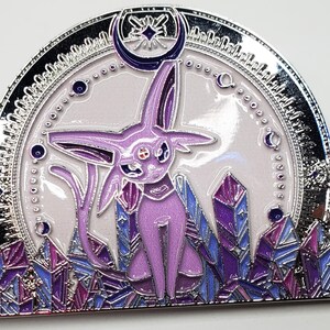 May include: A silver and purple enamel pin featuring a purple and pink Pokemon character with a crescent moon and stars above it. The character is surrounded by purple and blue crystals.