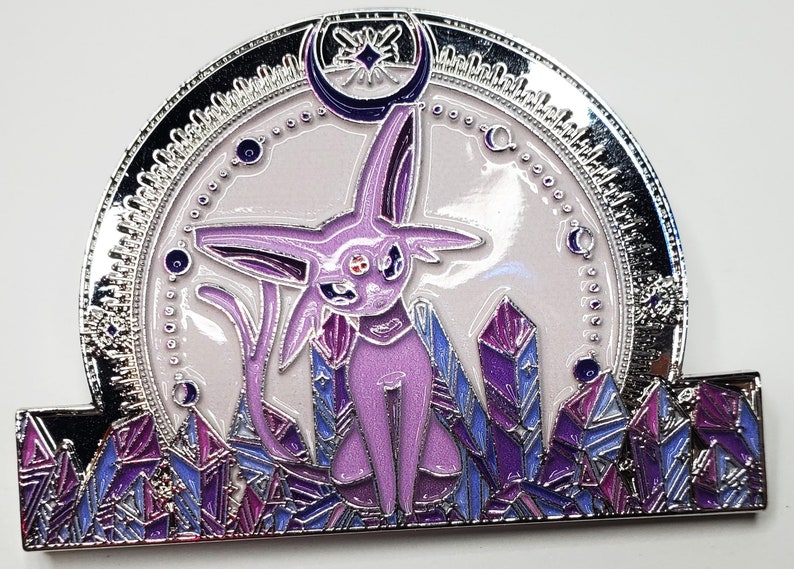 May include: A silver and purple enamel pin featuring a purple and pink Pokemon character with a crescent moon and stars above it. The character is surrounded by purple and blue crystals.