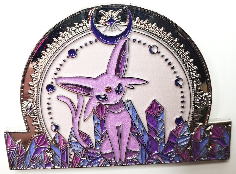 May include: A silver and purple enamel pin featuring a purple and pink Pokemon character, known as Espeon, sitting on a bed of purple crystals. A crescent moon with a star is above the character.