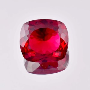 Puede incluir: Una piedra preciosa de color rojo intenso con talla cojín. La piedra preciosa está facetada y tiene un alto pulido.