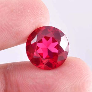 Puede incluir: Una piedra preciosa redonda roja con un corte en forma de estrella. La piedra preciosa está facetada y tiene un alto brillo.