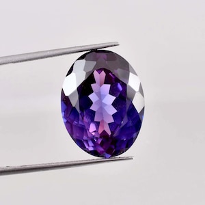 May include: An oval-shaped gemstone in shades of purple, held by tweezers. The gem's cut creates multiple facets that reflect light, showing a range of colours from dark violet to lighter purples and pinks. The background is plain white.