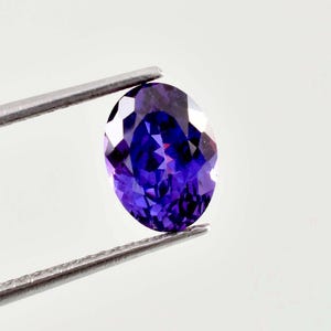 Può includere: Una pietra preziosa dal taglio ovale con un colore viola profondo e vibrante e una superficie sfaccettata che riflette la luce. La pietra è tenuta da una pinzetta in metallo color argento, che ne mostra la chiarezza e la finitura lucida.