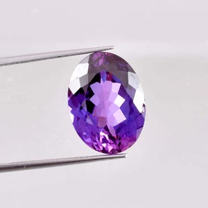 以下が含まれることがあります： ファセットカットされた大きな楕円形の紫色の宝石。宝石は白い背景に対してピンセットで保持されています。