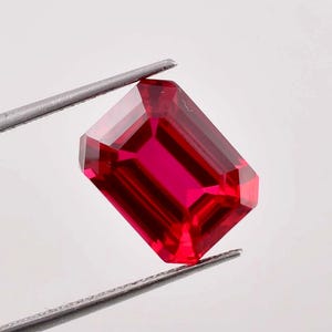 Puede incluir: Una gema roja vibrante de talla esmeralda sostenida por unas pinzas. La gema es de un color rojo intenso y rico y tiene un alto nivel de claridad.