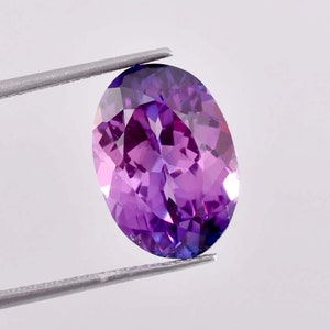 May include: A vibrant purple oval-shaped gemstone with a faceted cut, held by tweezers against a white background.