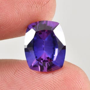 May include: A cushion-cut gemstone in a deep purple shade, held between fingers. The gem's facets catch the light, producing a vibrant display of colour. The stone is rectangular with rounded corners, highlighting its polished surface.