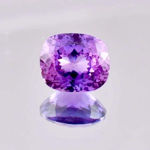 Può includere: Una gemma viola vibrante con taglio a cuscino. La gemma ha una tonalità viola chiaro con accenti viola più scuri. La gemma riflette la luce su una superficie bianca.