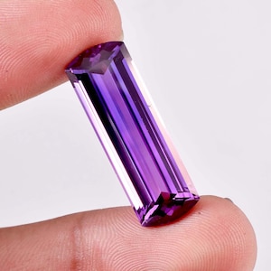 Peut inclure: Une pierre précieuse d'améthyste rectangulaire et facettée, d'un violet profond. La gemme est tenue entre deux doigts, mettant en valeur ses surfaces polies et ses facettes internes. La couleur de la pierre passe du violet foncé au violet clair.