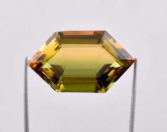 May include: A faceted, octagonal, yellow-green gemstone with a golden hue. The gemstone is held in place by two metal prongs.