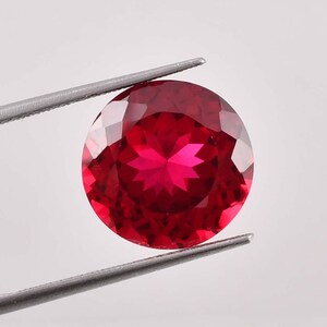 Puede incluir: Una piedra preciosa de color rojo intenso con un patrón en forma de estrella en el centro. La piedra preciosa es redonda y tiene un corte facetado.