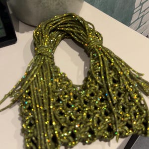 May include: A small handbag crafted from olive green cord with a shimmering, glittery finish. The bag features an open-weave macrame pattern and two integrated handles formed by the continuation of the knotted cord structure.