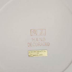 May include: A close-up of a white ceramic plate with a slightly curved rim. The plate has a brown stamp with Japanese characters and the words "HAND DECORATED". A gold sticker reads "CARNART IMPORTS, INC 1982 MADE IN JAPAN".