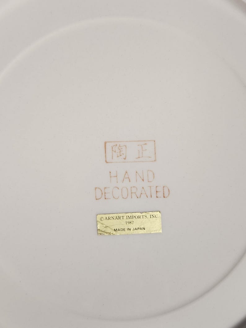 May include: A close-up of a white ceramic plate with a slightly curved rim. The plate has a brown stamp with Japanese characters and the words "HAND DECORATED". A gold sticker reads "CARNART IMPORTS, INC 1982 MADE IN JAPAN".