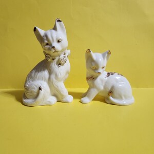 May include: Two white porcelain cat figurines with gold accents. The larger cat is sitting with a gold collar and a rose design. The smaller cat is lying down with a gold collar and a floral design.