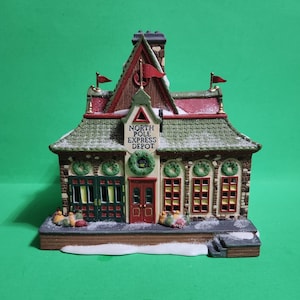May include: A decorative Christmas village building with the text "North Pole Express Depot". The building has a red roof, green shingles, and a stone facade. It features wreaths, flags, and snow accents, creating a festive holiday scene.