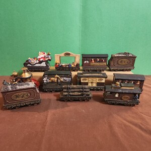 May include: A collection of miniature train cars, each with intricate details. The cars are primarily brown with gold accents, depicting various scenes and cargo. One car features Santa Claus in a sleigh. The set includes a variety of train cars.