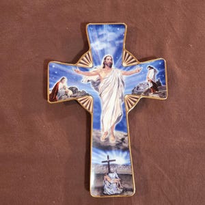 May include: A ceramic cross with a gold trim, featuring a religious scene. The cross depicts Jesus with outstretched arms, surrounded by other figures and a blue sky background.