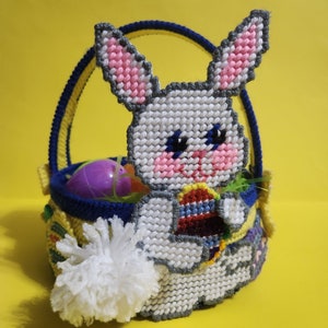 May include: A white bunny-shaped Easter basket with a blue handle and a white pom-pom tail. The basket is filled with colorful Easter eggs and green grass.