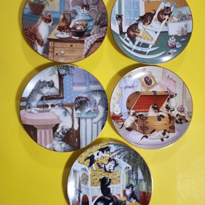 May include: Five decorative plates featuring cat illustrations. Each plate depicts cats in various domestic settings, including kitchens and living rooms. The plates have gold rims and showcase detailed, colorful artwork of felines.