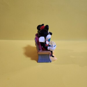 Pu&ograve; includere: Una statuetta in ceramica di Topolino e Minnie al cinema. Topolino indossa un papillon rosso e Minnie un cappello rosso. Sono seduti su una sedia viola con una base blu, tenendo in mano dei popcorn.