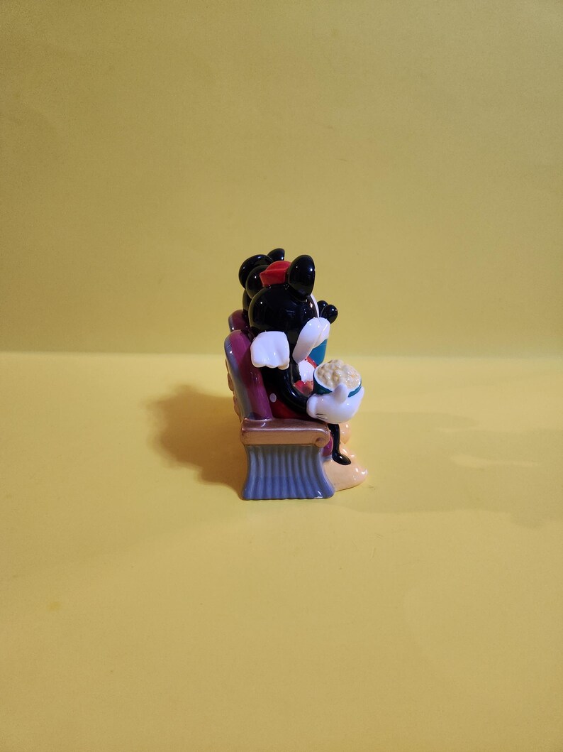 Pu&ograve; includere: Una statuetta in ceramica di Topolino e Minnie al cinema. Topolino indossa un papillon rosso e Minnie un cappello rosso. Sono seduti su una sedia viola con una base blu, tenendo in mano dei popcorn.