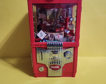 May include: Red Coca-Cola themed music box with a glass front. The music box has a coin slot and a small window with a miniature scene inside. The front of the music box has the Coca-Cola logo and the words "Insert Coin Here".