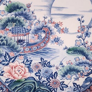 May include: A ceramic plate with an Asian-inspired design in blue and white. The plate depicts a boat, flowers, and a landscape scene, with a large pink peony as a focal point. The rim has a red and white patterned border. The design is highly detailed.