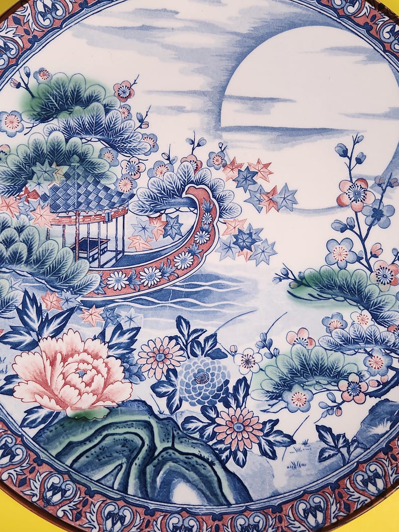 May include: A ceramic plate with an Asian-inspired design in blue and white. The plate depicts a boat, flowers, and a landscape scene, with a large pink peony as a focal point. The rim has a red and white patterned border. The design is highly detailed.
