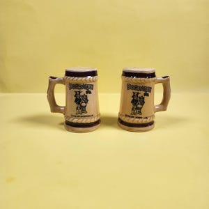 May include: Two ceramic salt and pepper shakers shaped like miniature beer steins. Each stein is tan with a dark brown rim and handle. The text "Dogpatch" is printed on the front, along with a graphic of two cartoon characters. The base of each stein has additional text.