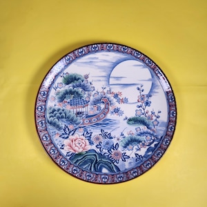 May include: A round decorative plate with an Asian-inspired design. The plate features a pagoda, flowers, and a large moon in blue, pink, and green. The rim has a patterned border. The plate's diameter is approximately 25 cm.