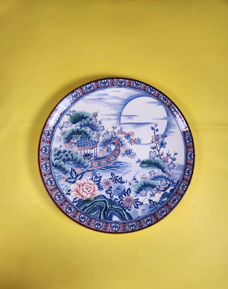May include: A round decorative plate with an Asian-inspired design. The plate features a pagoda, flowers, and a large moon in blue, pink, and green. The rim has a patterned border. The plate's diameter is approximately 25 cm.