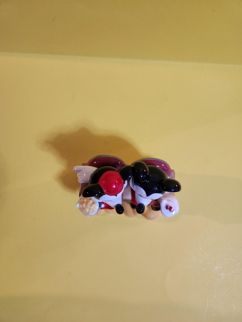 Pu&ograve; includere: Un paio di statuette di orche in ceramica, una con una palla rossa sulla testa e l'altra con una testa nera. Le orche sono nere e bianche con accenti rossi e bianchi. Sono appoggiate su una base rossa e viola.