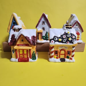 May include: Set of five ceramic Christmas village houses with snow-covered roofs. The houses are painted in various colors, including red, yellow, blue, and white. The houses have different designs, including a schoolhouse, a candy house, and a toy shop. The houses are sitting on a wooden surface.