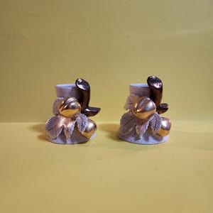 May include: Two ceramic candle holders with a speckled white and gold finish. Each holder features a gold-colored fruit and leaf design at the base, with a dark brown, curved accent at the top. The background is a solid yellow.