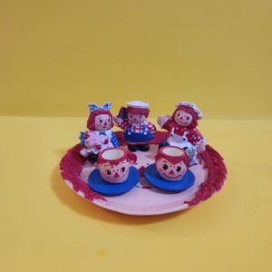 May include: A miniature tea set featuring Raggedy Ann and Andy dolls. The set includes a pink plate with a red trim, two blue saucers with matching cups, and three doll figurines. The dolls are dressed in red, white, and blue outfits.